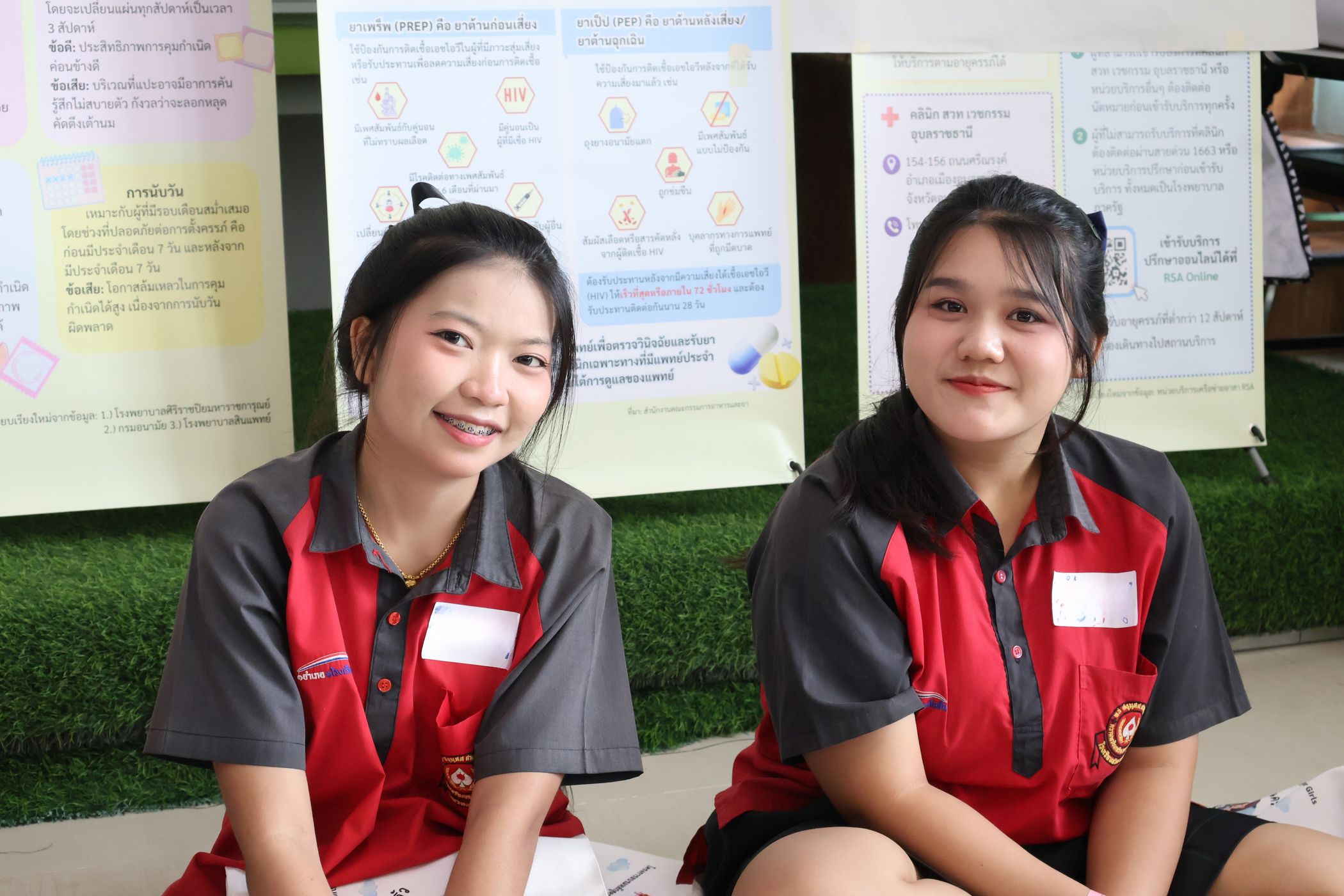 Two smiling students in red and gray school uniforms sit in front of health education posters at a Better Me for Girls workshop in Ubon Ratchathani, Thailand.