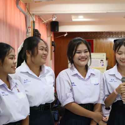 Four smiling students in white school uniforms stand together as one speaks into a microphone during a Better Me for Girls workshop in Thailand.